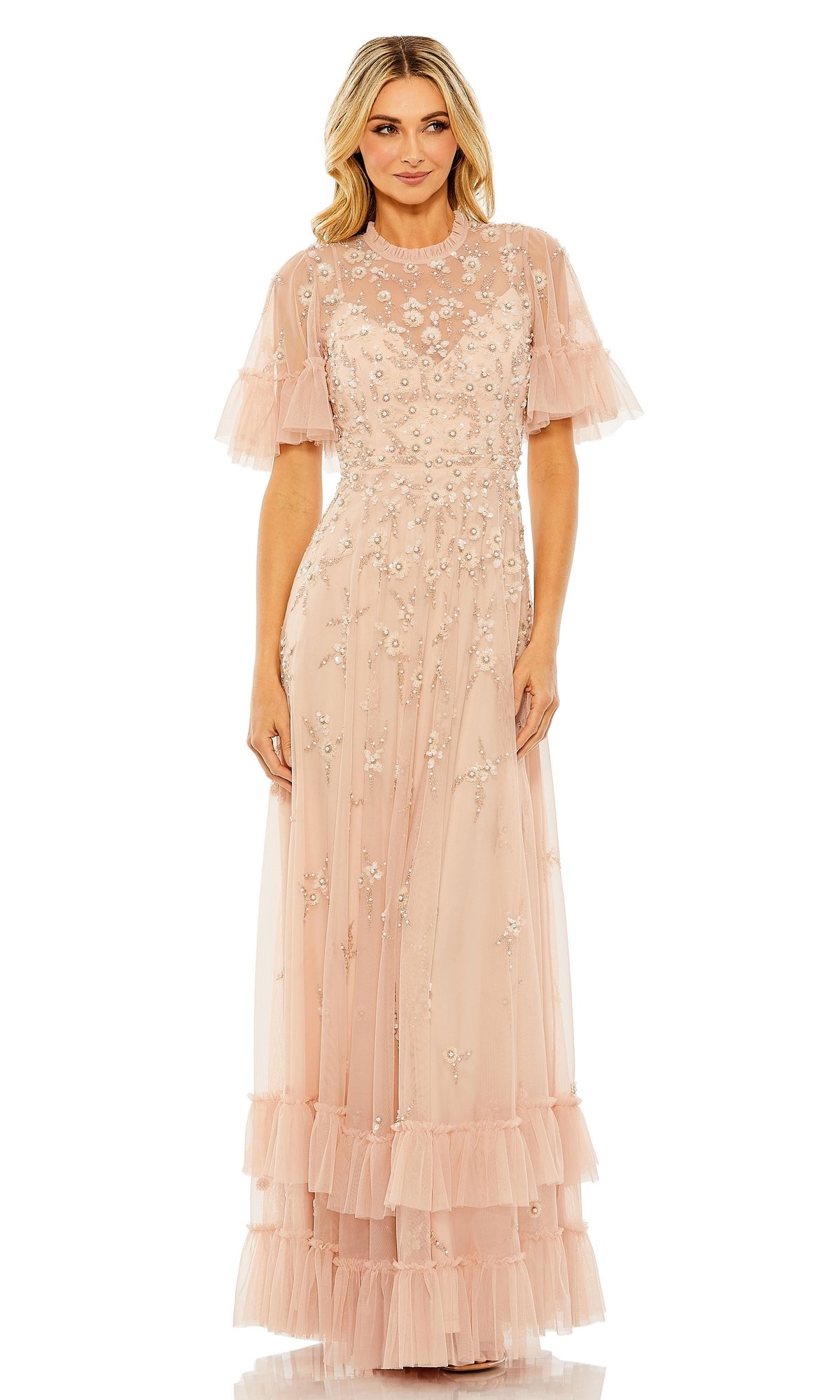 A woman wearing the Mac Duggal 9199 Short Sleeve Beaded Formal Gown—a floor-length blush pink dress with floral embroidery, sheer overlay, and ruffled hem—stands facing forward.
